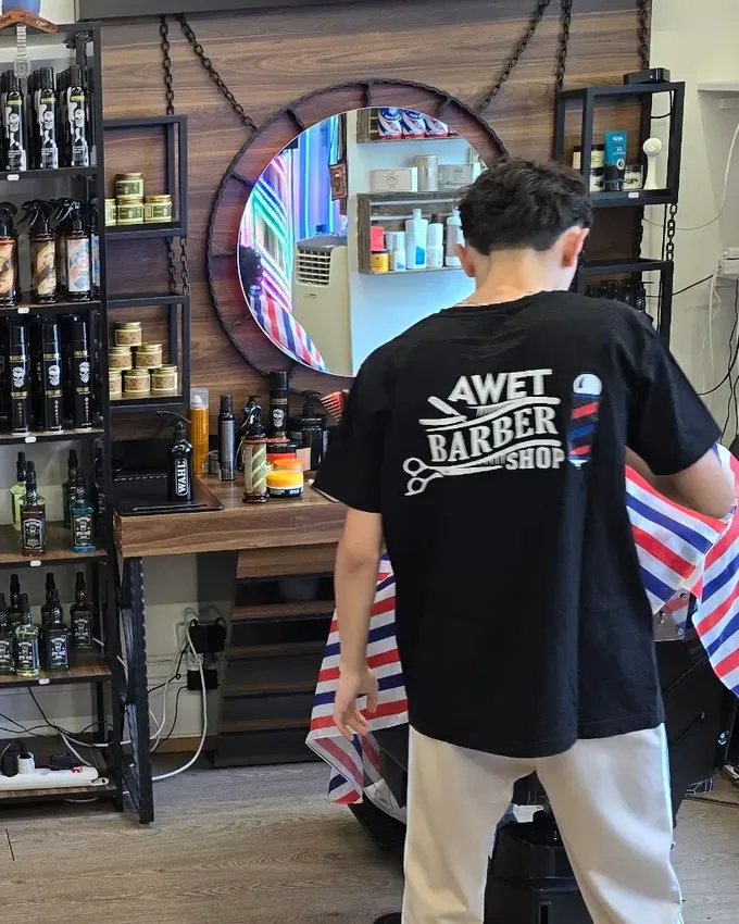 Barber im schwarzen Awet-Barber-Shop-Shirt vor dem runden Spiegel und den Regalen mit Pflegeprodukten