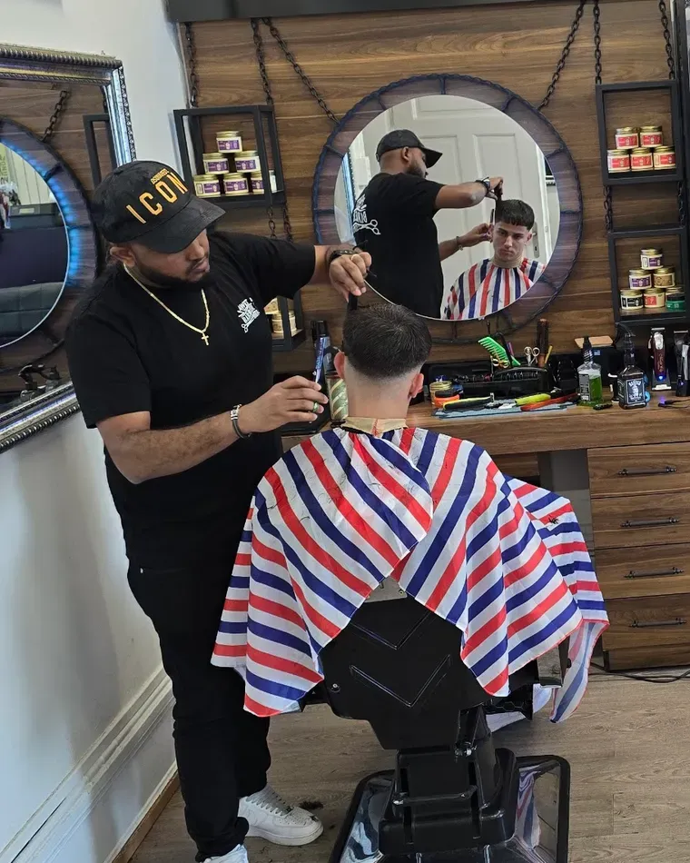 Ein Barber schneidet einem Kunden im gestreiften Umhang die Haare, im runden Spiegel spiegelt sich die Szene
